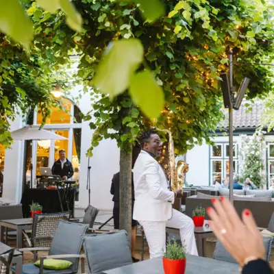 Événements sur la terrasse des tilleuls Un homme en costume blanc joue du saxophone sous un arbre.
